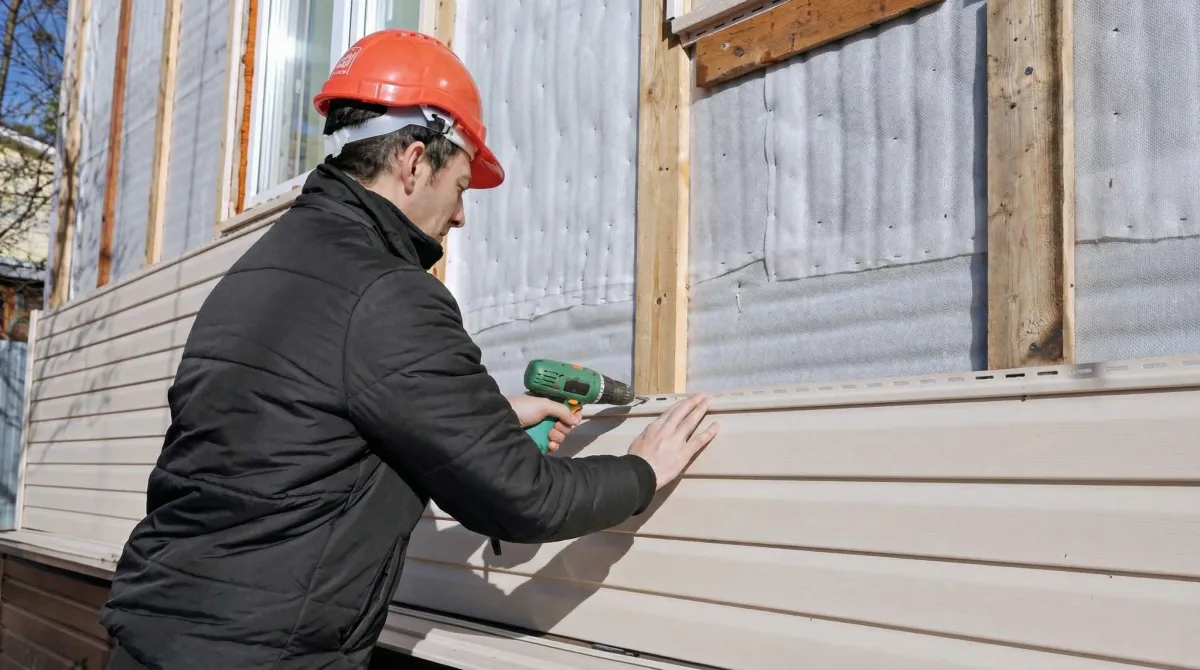 Siding replacement professional using a power drill to install new tan vinyl siding on a home exterior in Kansas City, KS